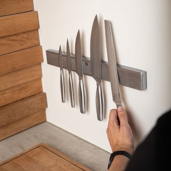 Een hand pakt een van de vijf messen van een magnetische strip die aan de muur hangt. De messen hebben zilverkleurige handvatten en roestvrijstalen lemmeten.