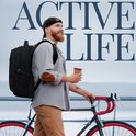 Man met baard, pet en rugzak, lopend met een fiets en een koffiebeker. Tekst 'ACTIVE LIFE'.