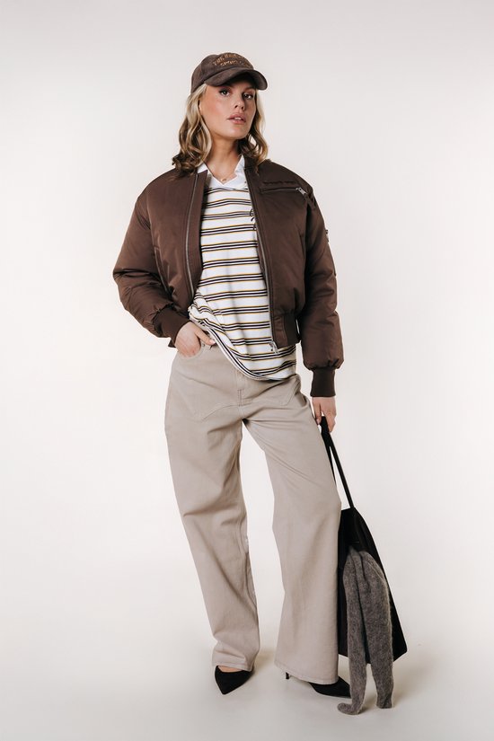 Model in bruine bomberjack, gestreept shirt en wijde beige broek, met tas.
