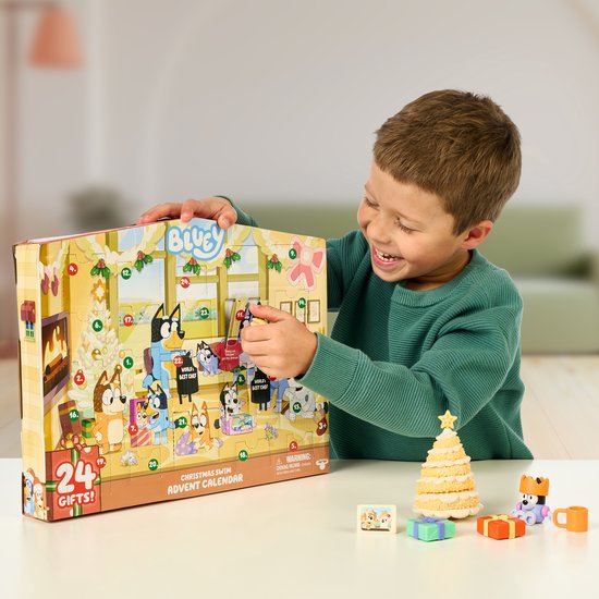 Jongen opent Bluey Kerst zwem adventskalender met 24 vakjes. Een kerstboom en speelgoed liggen naast de kalender op tafel.