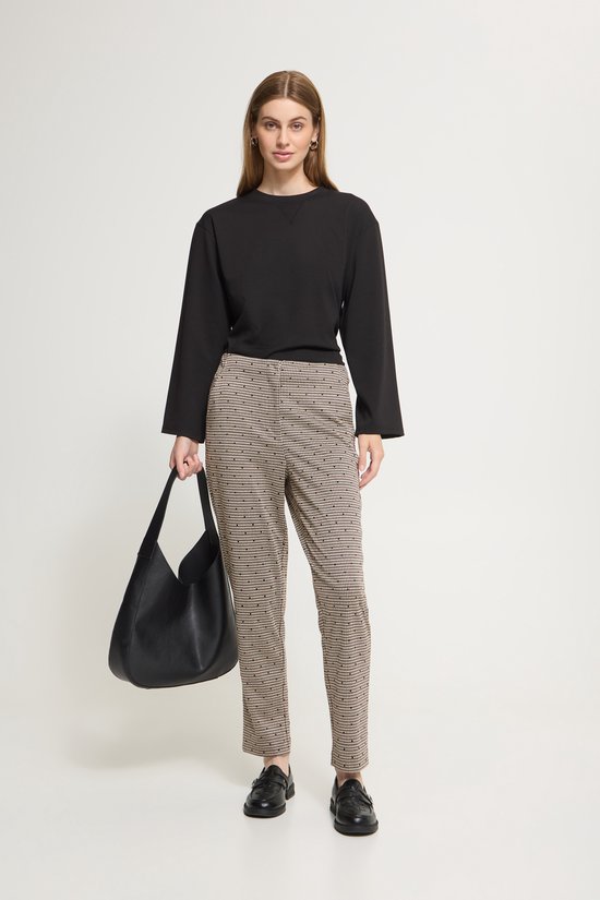 Model met een beige broek met ruitpatroon en zwarte stippen, zwart shirt, zwarte tas.