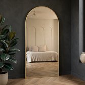 Boogspiegel met gouden rand, staand op een houten vloer, weerspiegelt een bed en muurdecoratie.