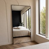 Grote rechthoekige spiegel met zwarte lijst, weerspiegelt een slaapkamer met een bed en grote ramen met uitzicht op groen.