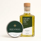 Glazen fles met druivenpitolie en rond blikje bijenwas, beide met donkergroene etiketten en gouden accenten.