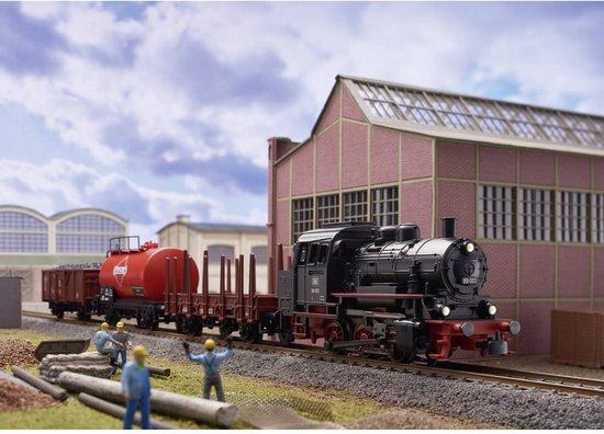 Zwarte stoomlocomotief met rode details, met goederenwagons waaronder een rode tankwagon, bij een fabriek.