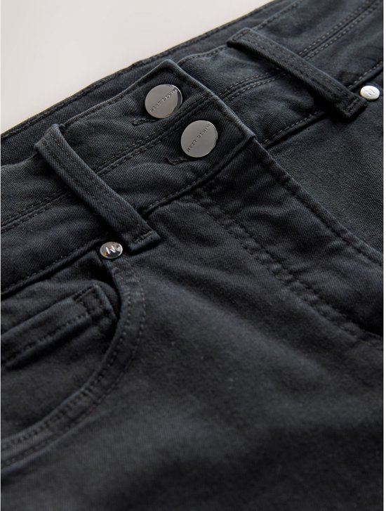 Detailopname van de tailleband van een zwarte jeans met twee zilverkleurige 'NEXT DENIM' knopen.