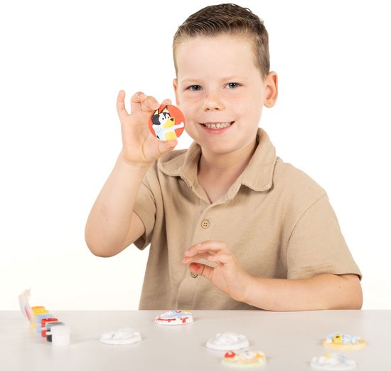 Jongen in beige poloshirt houdt een handgemaakte Bluey gipsfiguur omhoog, omringd door verfpotjes en gipsfiguren.