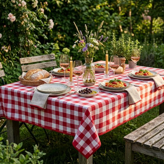 Une nappe à motif à carreaux rouge et blanc, dressée avec des assiettes de pâtes, des olives, du pain et des fleurs dans un jardin.