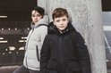 Twee jongens die poseren in winterjassen, de ene in het zwart, de andere in het lichtgrijs, leunend tegen een betonnen pilaar.
