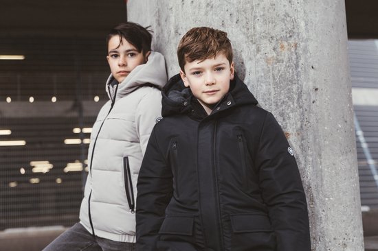 Twee jongens die poseren in winterjassen, de ene in het zwart, de andere in het lichtgrijs, leunend tegen een betonnen pilaar.