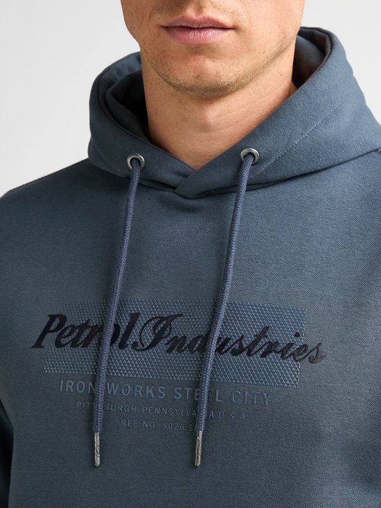 Close-up van een blauwe hoodie met Petrol Industries-tekst en trekkoorden met metalen uiteinden.