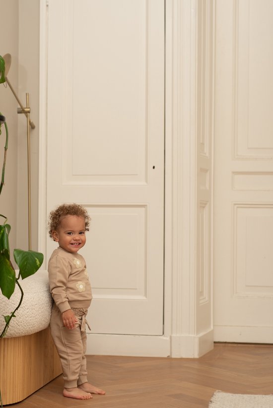 Een lachend jong kind met krullend haar in een lichtbruine outfit met bloemenprint.