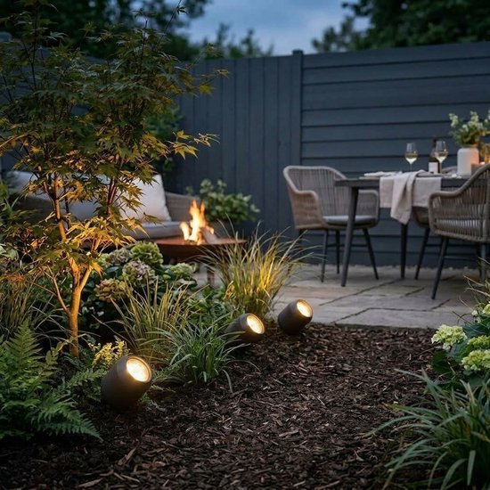 Drie donkergrijze grondspots belichten een tuinpad met mulch, omringd door groen en een zitje met vuurkorf.