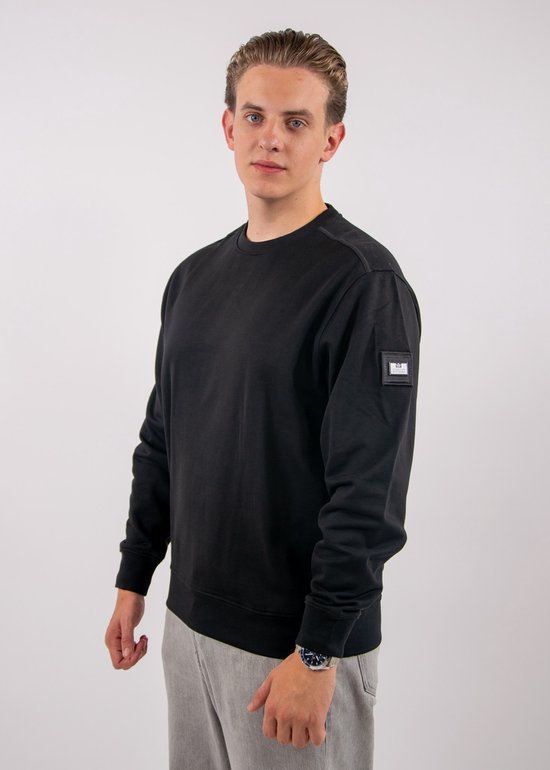 Model draagt een zwarte sweater met ronde hals en een duidelijk zichtbare logopatch op de linkermouw.