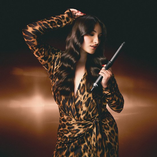 Model met lang, golvend haar en panterprint jurk, houdt de ghd Chronos Curve Classic krultang vast.