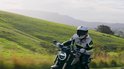Motorrijder met een wit helm en grijs/groene jas op een motor, groen landschap.