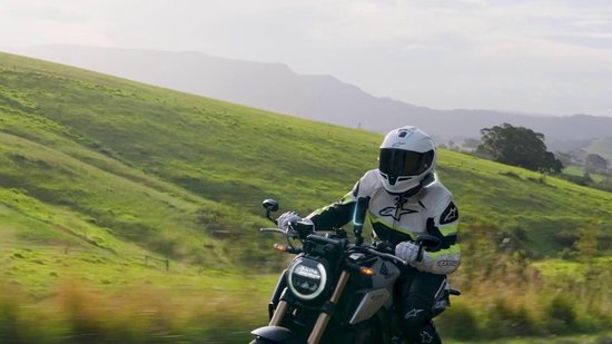 Motorrijder met een wit helm en grijs/groene jas op een motor, groen landschap.