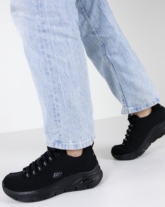 Een persoon draagt zwarte Skechers Arch Fit sneakers met een lichtblauwe jeans.