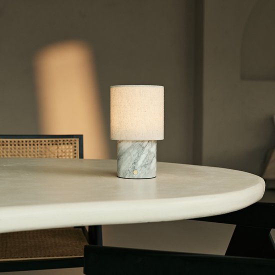 Marmeren tafellamp met linnen kap staat op een witte tafel naast een stoel met rieten zitting.
