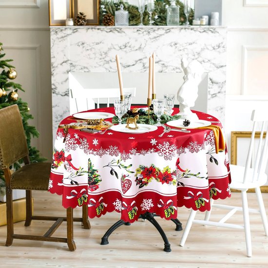 Gedekte ronde kersttafel met rood tafelkleed, witte sneeuwvlokken en rand met kerstbomen en bloemen. Kerstsfeer met kerstboom en open haard.