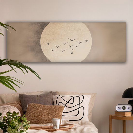 Een langwerpig canvas met een grote, licht beige cirkel tegen een beige achtergrond. Een groepje donkere vogels vliegt in formatie voor de cirkel. Hangt boven een bed met kussens en een nachtkastje.
