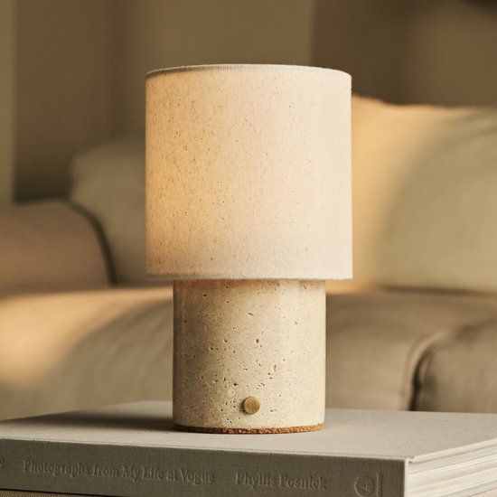 Cilindrische tafellamp met beige linnenkap en travertijnen voet, staand op een boek.