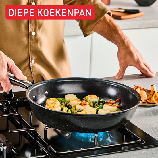 Een diepe koekenpan met een vis, groenten en paddenstoelen op een gasfornuis, vastgehouden door een persoon.