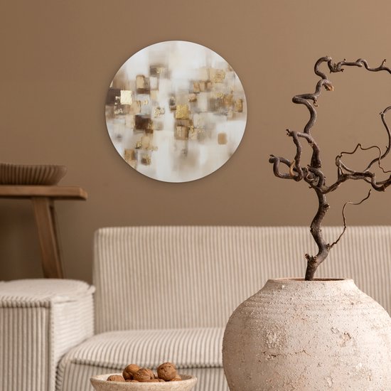 Een ronde muurcirkel met abstracte gouden en bruine vierkanten hangt aan een taupekleurige muur boven een witte bank.