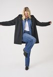 Vrolijke vrouw met blonde haren, draagt een zwarte lange jas met capuchon, denim overhemd en jeans.