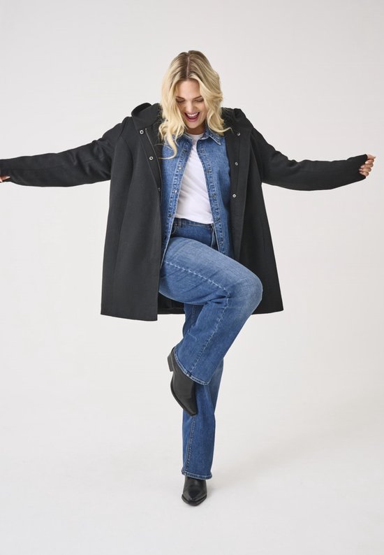 Vrolijke vrouw met blonde haren, draagt een zwarte lange jas met capuchon, denim overhemd en jeans.