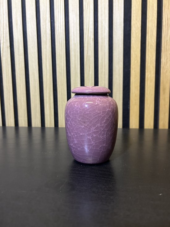 Kleine paarse urn met craquelé-afwerking en een hartvormige deksel, staand op een zwarte tafel.