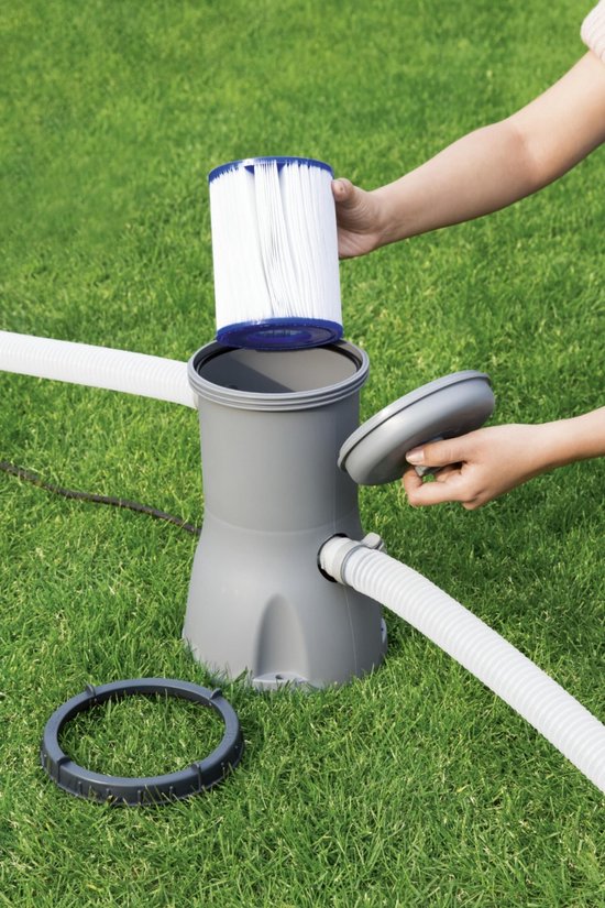 Een persoon vervangt een wit pliss filter met blauwe uiteinden in een grijze zwembadpomp op groen gras.