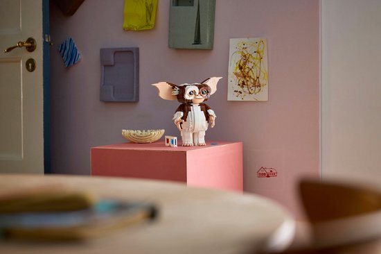 Een bruin-witte LEGO Ideas Gizmo figuur met grote oren staat op een roze kubus. Een rieten kom staat ernaast.