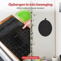 Een hand legt een zwarte ronde inductiebeschermer met ruitpatroon op een aanrecht, daarnaast hangt er één aan de muur.