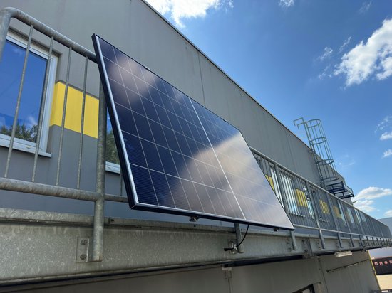 Zwart zonnepaneel met rasterpatroon, gemonteerd op een railing aan de muur.