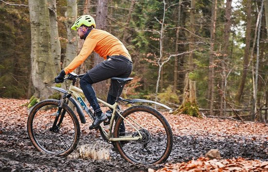 Een persoon op een fiets rijdt door een modderig bos met spatborden.