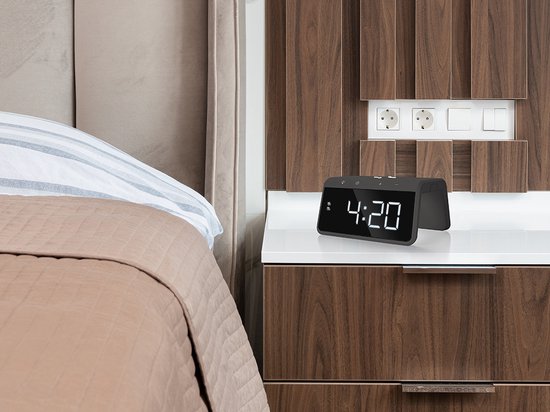 Zwarte digitale wekker met groot display, op een witte nachtkastje naast een bed.