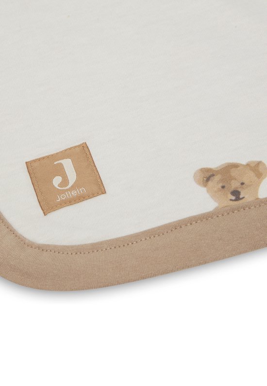 Détail du coin d'une couverture bébé blanche à imprimé ours en peluche, avec un bord marron et une étiquette 'Jollein'.