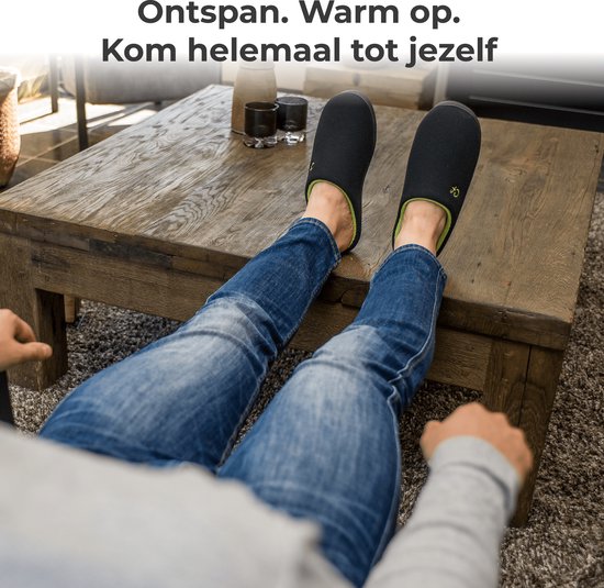 Man met spijkerbroek ontspant met voeten omhoog op een salontafel, draagt zwarte pantoffels met groene voering.