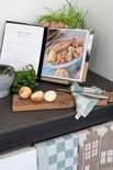 Keuken met kookboek, uien, kruiden en gebreide theedoeken met blokjes- en huisjespatroon.