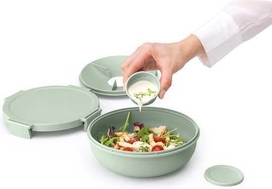 Une main ajoute de la vinaigrette d'un petit récipient vert jade à une salade dans une boîte à salade.