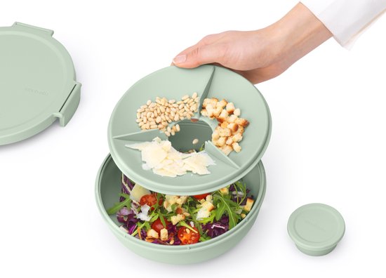Une main ouvre le couvercle segmenté d'une boîte à salade verte jade, contenant des pignons, des croûtons et du fromage.