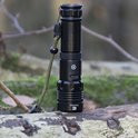 Een zwarte Exortus LED-zaklamp met een groen koord, staand op een boomstronk in het bos.