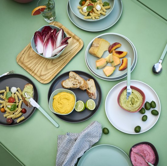 Een tafel met een zomerse maaltijd: salades, brood, dips en fruit. Verschillende kommen en borden staan verspreid.