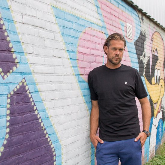 Man met zwarte T-shirt met korte mouwen en blauwe joggingbroek, leunend tegen muur met graffiti.