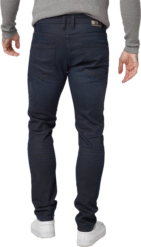 Een man met tatoeages draagt een grijze longsleeve en een donkerblauwe slim fit jeans, van achteren gezien.