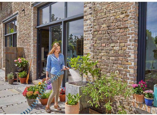 Vrouw in blauw hemd en bruine broek besproeit planten met blauwe spiraalslang naast een bakstenen gebouw.