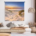 Wandkleed met zonsondergang boven duinen, hangend in een woonkamer boven een witte bank, met tekst over afwerking.