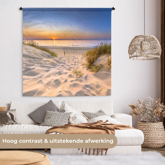 Wandkleed met zonsondergang boven duinen, hangend in een woonkamer boven een witte bank, met tekst over afwerking.