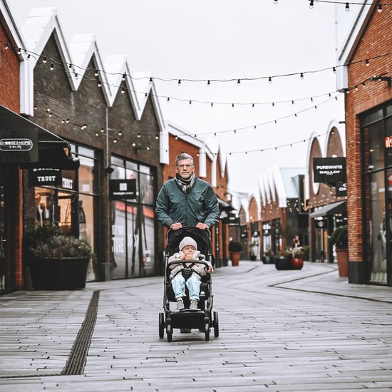 Man met grijze sjaal duwt een zwarte Deryan buggy met een baby erin, op een winkelstraat.